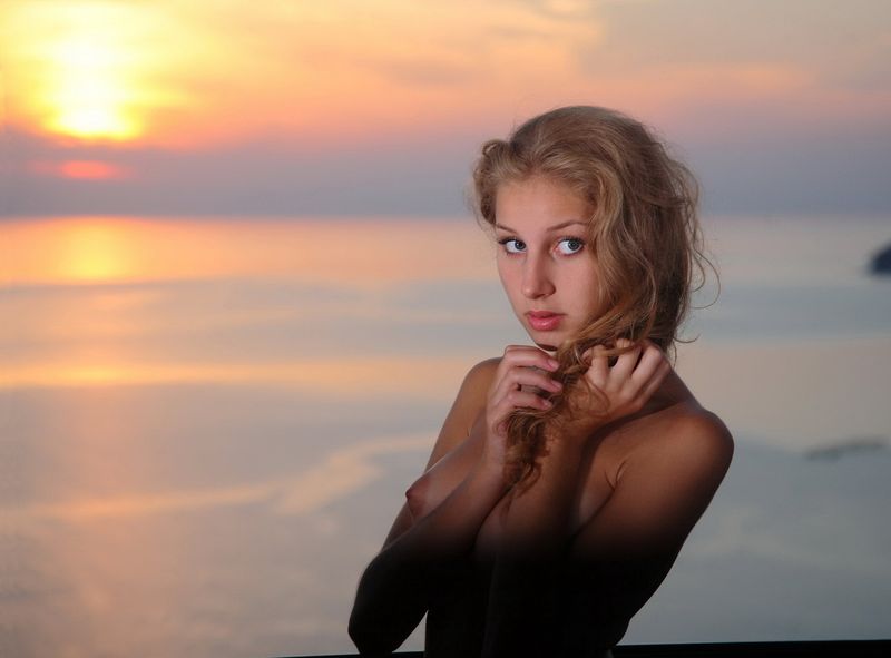cute young curly blonde girl with a necklace and pendant reveals her black chemise with stockings at home in the evening sunset