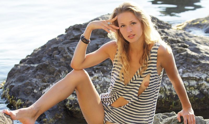 young blonde girl with tan lines reveals her black and white striped sleeveless tank top shirt on the rocky shore at the sea