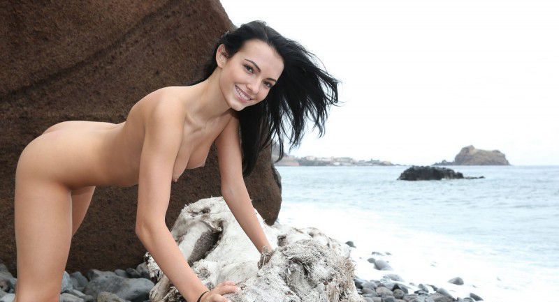 young black haired girl reveals her short jeans on the driftwood on the rocky shore at the sea