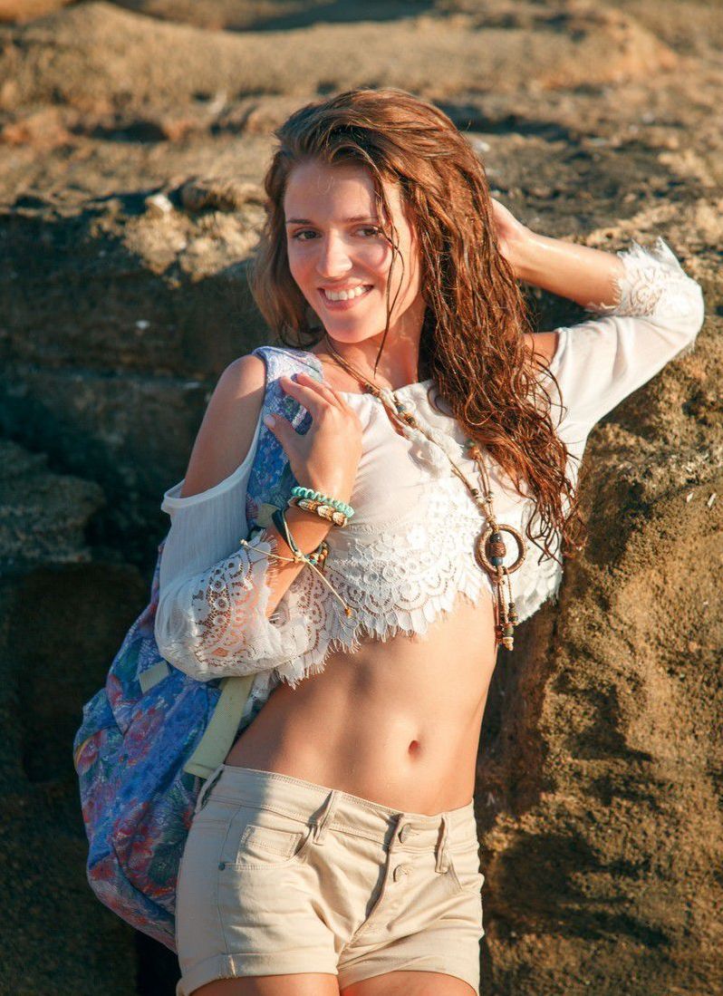 young curly red haired girl with a necklace reveals her white top and short jeans on the rocky shore at the sea